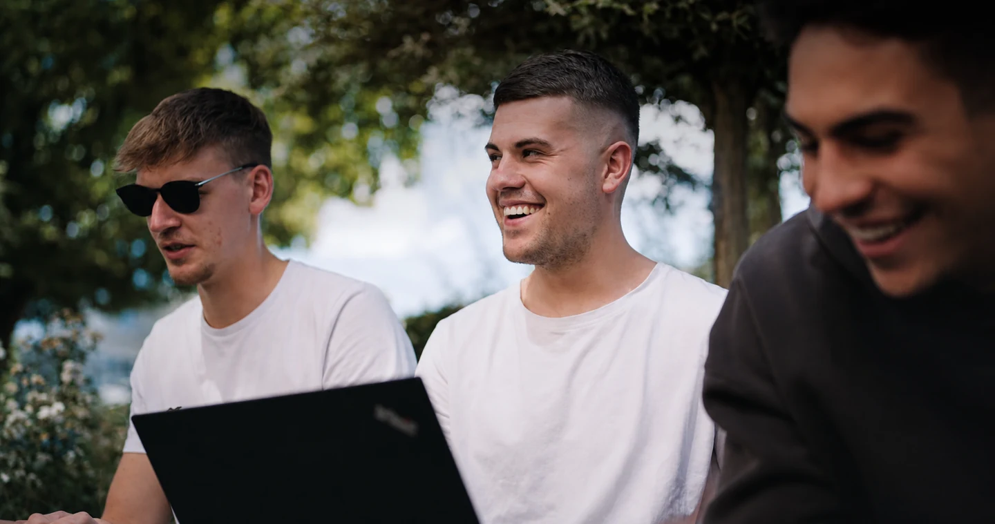Drei junge Studierende arbeiten draußen gemeinsam am Laptop und lachen