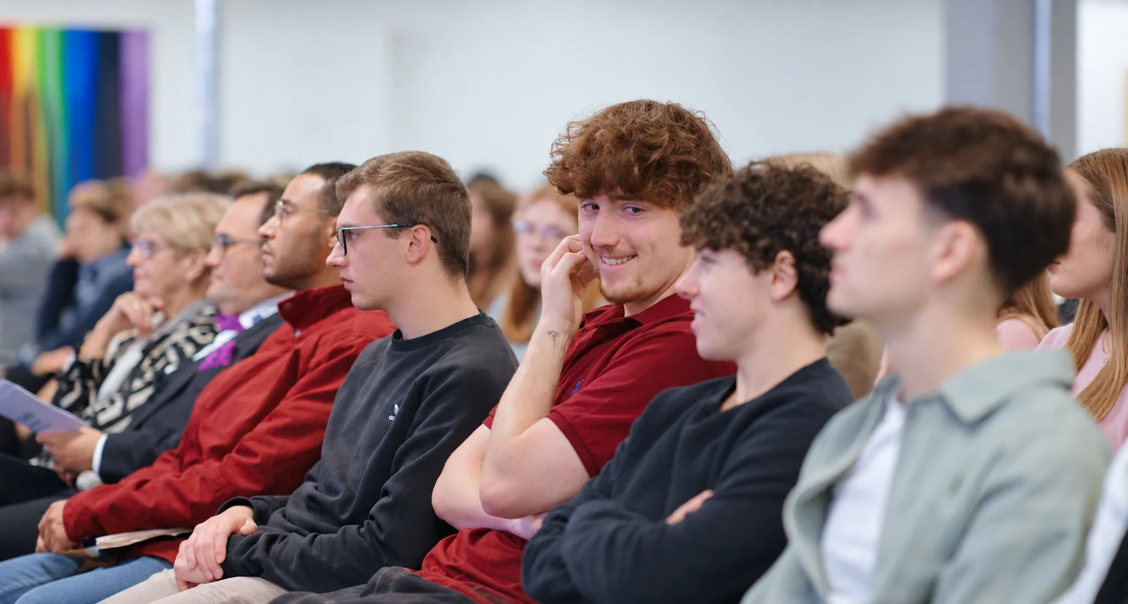 Junge Menschen sitzen bei einer Campusveranstaltung zusammen; ein Student im roten Poloshirt lächelt