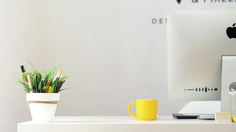 Ein heller Arbeitsplatz mit Computer, Pflanzen und einer gelben Tasse.